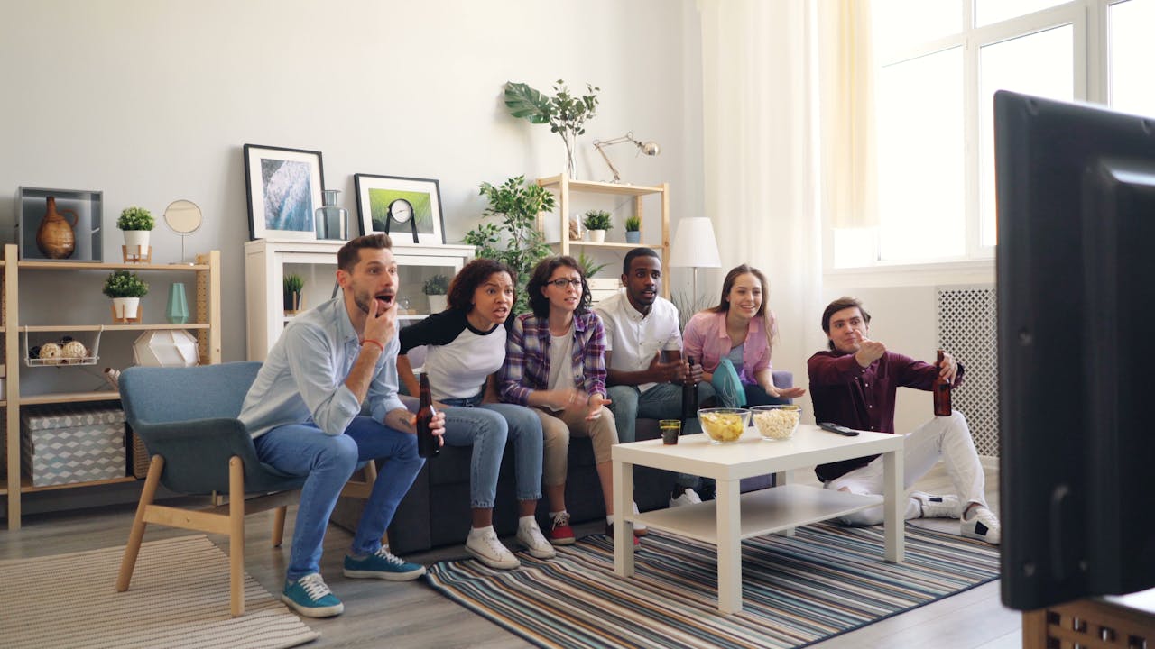 A diverse group of young adults excitedly watching a sports game indoors with snacks and drinks.