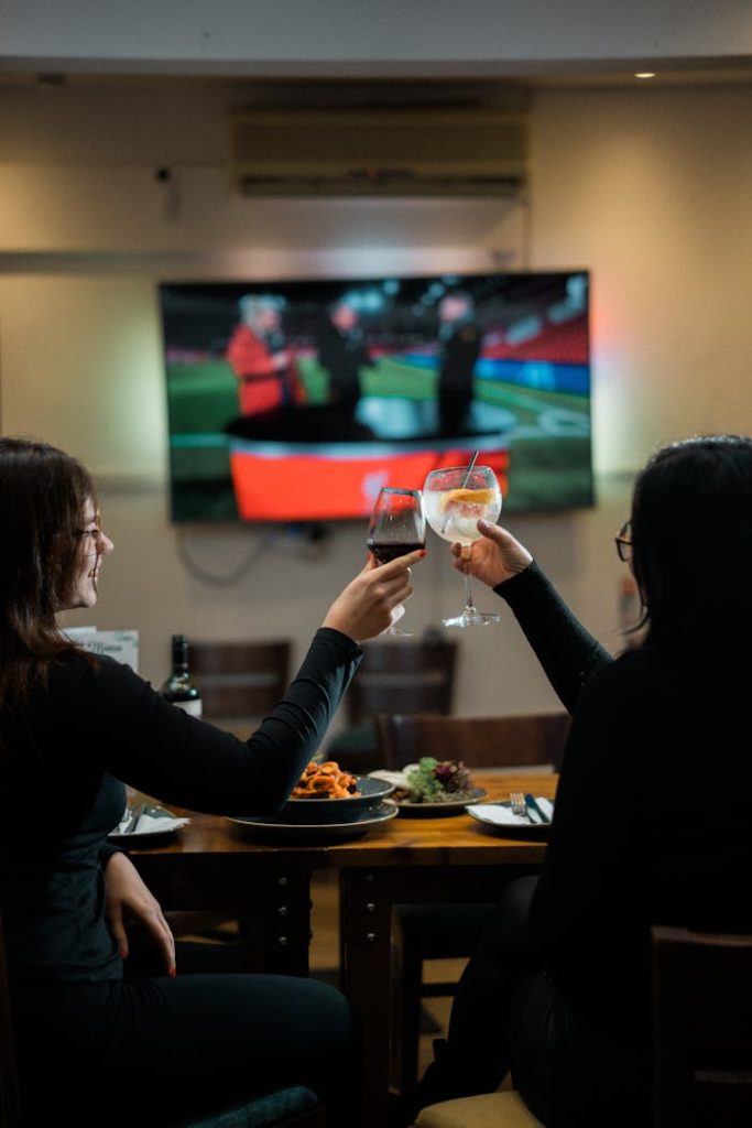 pexels-photo-31173327 Two women clinking wine glasses at a restaurant with a sports broadcast on TV in the background.